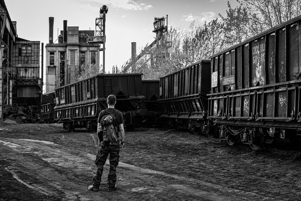 Schwarz-Weiß-Aufnahme eines Mannes vor einer Industrieruine. Im Vordergrund Eisenbahnwaggons.