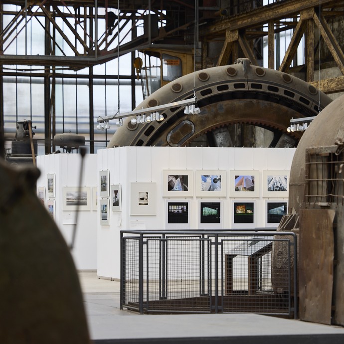 Blick auf eine Wand mit Bildern der Ausstellung, im Vorder- und Hintergrund sind Maschinen der Maschinenhalle zu sehen (vergrößerte Bildansicht wird geöffnet)