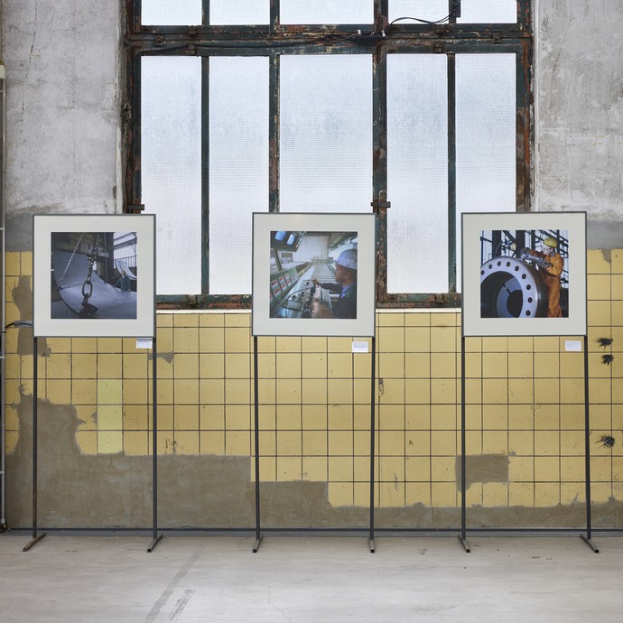 Blick auf eine gelb geflieste Wand von Industriefenstern durchbrochen, davorstehend sind drei Bilder der Ausstellung zu sehen (vergrößerte Bildansicht wird geöffnet)