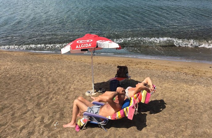 Ein Mann und eine Frau liegen in ihren Sonnenstühlen am Strand, auf der anderen Uferseite steht ein Stahl- und Eisenwerk. Montage: Sabine Korth