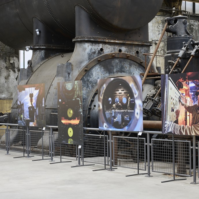 Blick in die Ausstellung und auf verschiedene Fotografien umgeben von historischen Maschinen der Maschinenhalle (vergrößerte Bildansicht wird geöffnet)