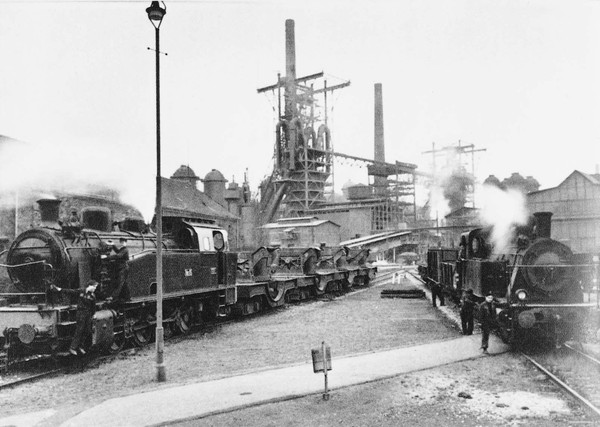 Historische Ansicht von zwei Dampflokomotiven, die über ein Hüttengelände fahren.